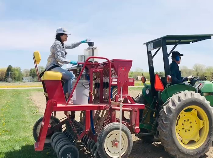 Woman seeding while sitting on the back of a tractor.